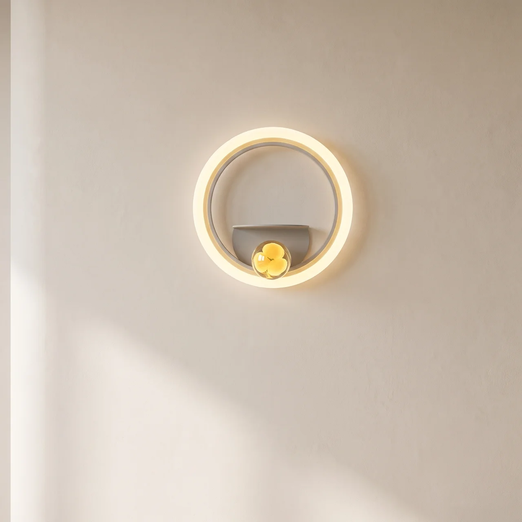A modern circular wall sconce light fixture with a glowing warm white LED ring, a gray inner arch, and a small yellow flower accent at the bottom center, mounted on a plain beige wall.