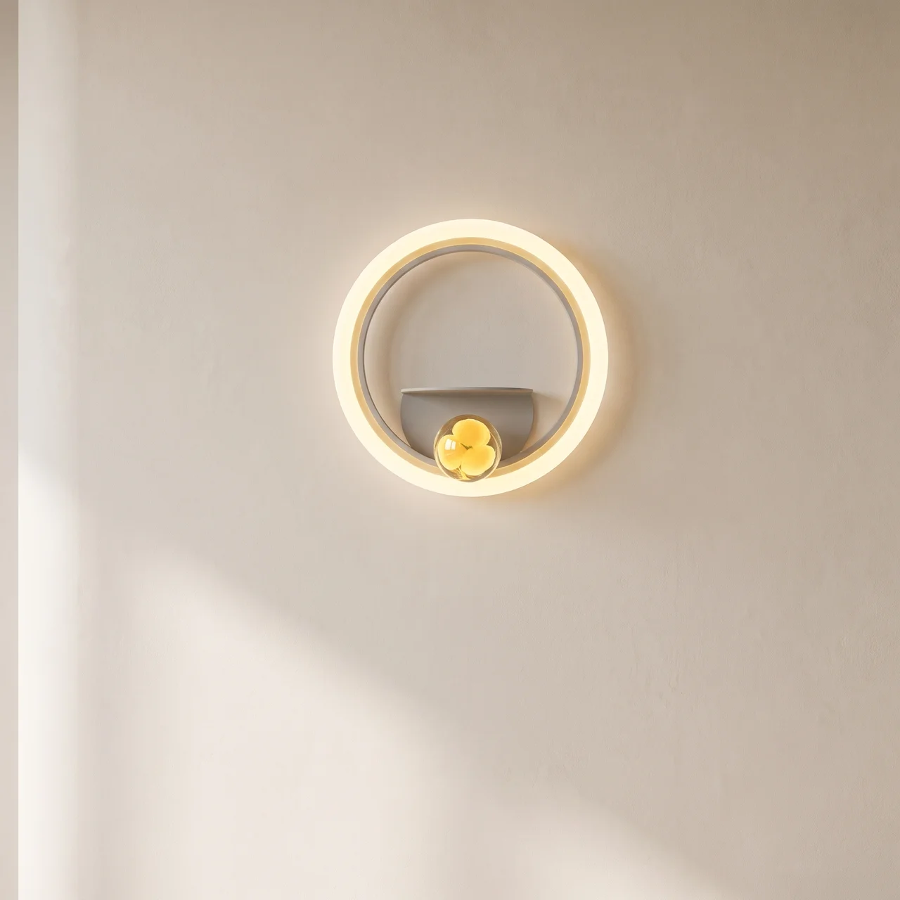 A modern circular wall sconce light fixture with a glowing warm white LED ring, a gray inner arch, and a small yellow flower accent at the bottom center, mounted on a plain beige wall.
