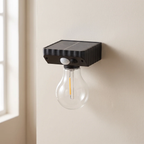 A black solar-powered outdoor wall light with a clear glass bulb, mounted on a beige wall next to a window.