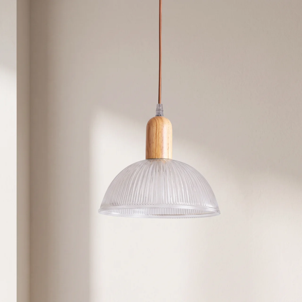 A modern pendant light with a clear ribbed glass dome shade, a light natural wood neck, and a brown cord, hanging against a warm beige wall.