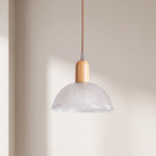 A modern pendant light with a clear ribbed glass dome shade, a light natural wood neck, and a brown cord, hanging against a warm beige wall.