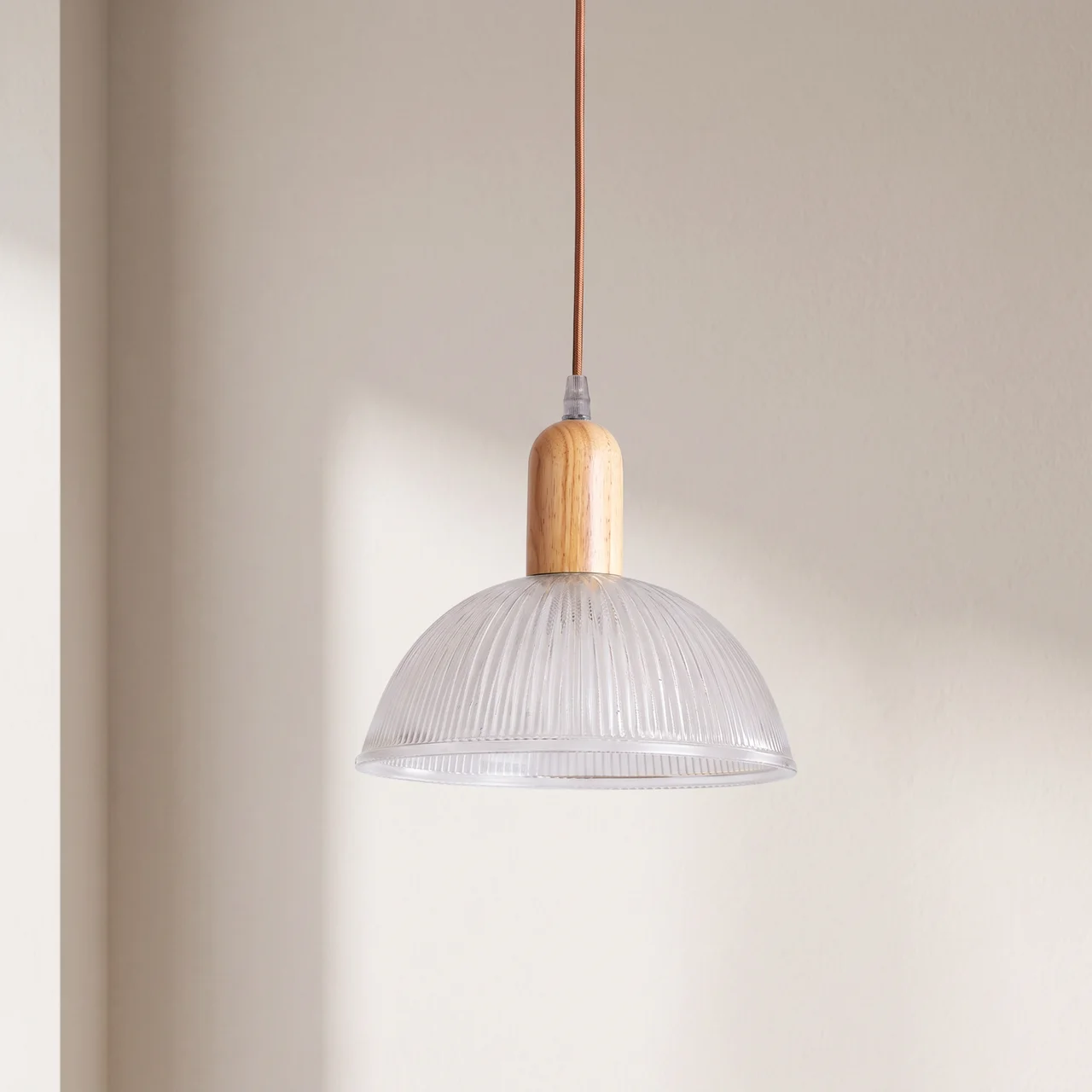 A modern pendant light with a clear ribbed glass dome shade, a light natural wood neck, and a brown cord, hanging against a warm beige wall.