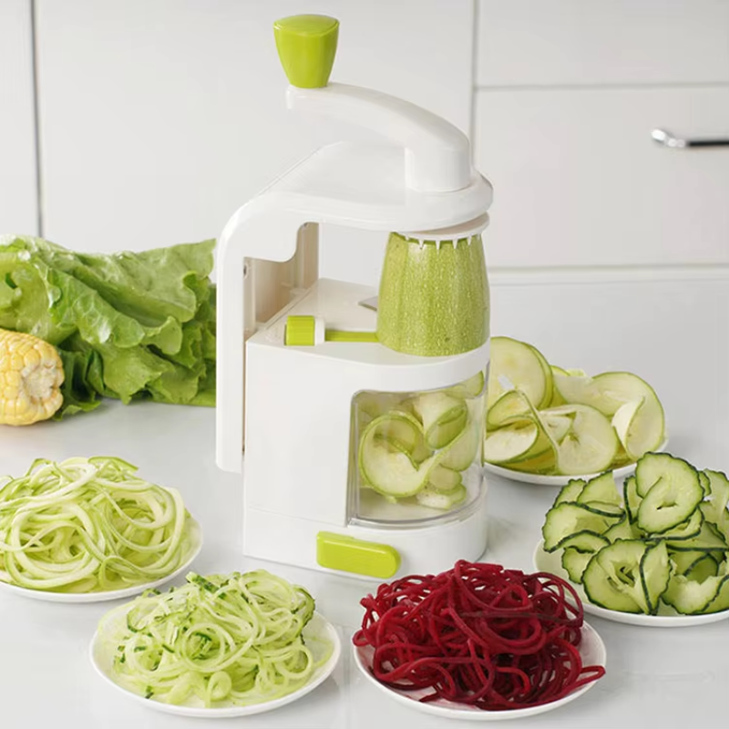 A white manual spiralizer with a green handle and green accents stands on a kitchen counter, with plates of sliced vegetables including zucchini, beet, and lettuce around it.