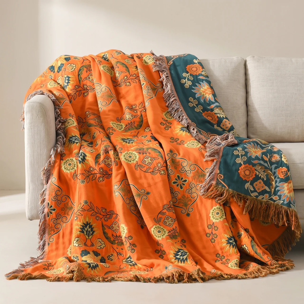 An orange patterned throw blanket with fringe and teal floral sections draped over a light beige sofa in a neutral indoor setting.