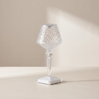 A clear crystal table lamp with a textured conical shade, a faceted stem, and a square base, set against a warm beige background with soft sunlight casting shadows.