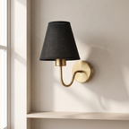 A modern wall sconce with a tapered black fabric shade and a curved brass arm mounted on a beige wall next to a window.