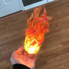 A hand holding a small flame-shaped decorative object with glowing orange and yellow faux flames, photographed over a wood floor.