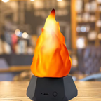 A tabletop flame-shaped lamp with a glowing orange and yellow faux flame on a dark gray geometric base, set against a blurred indoor background.
