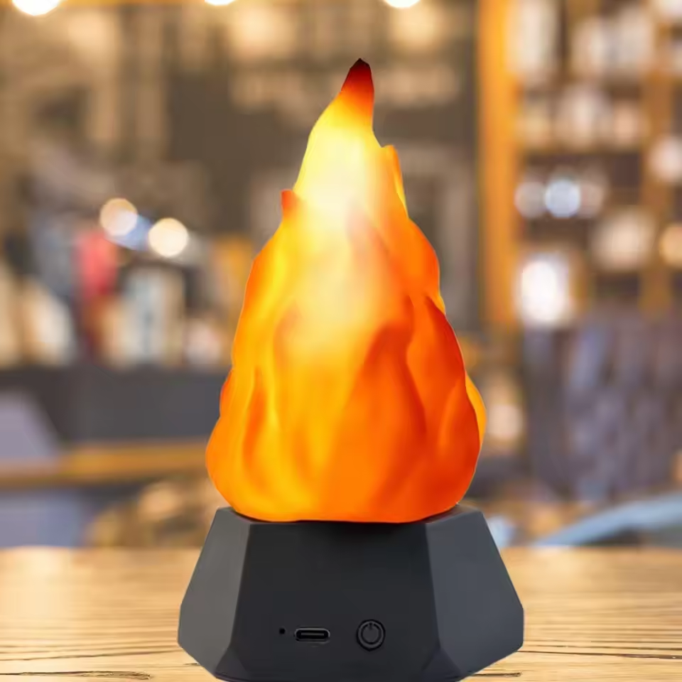 A tabletop flame-shaped lamp with a glowing orange and yellow faux flame on a dark gray geometric base, set against a blurred indoor background.