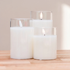 Three white pillar candles in clear glass holders of varying heights, arranged on a light wood surface against a soft beige background.