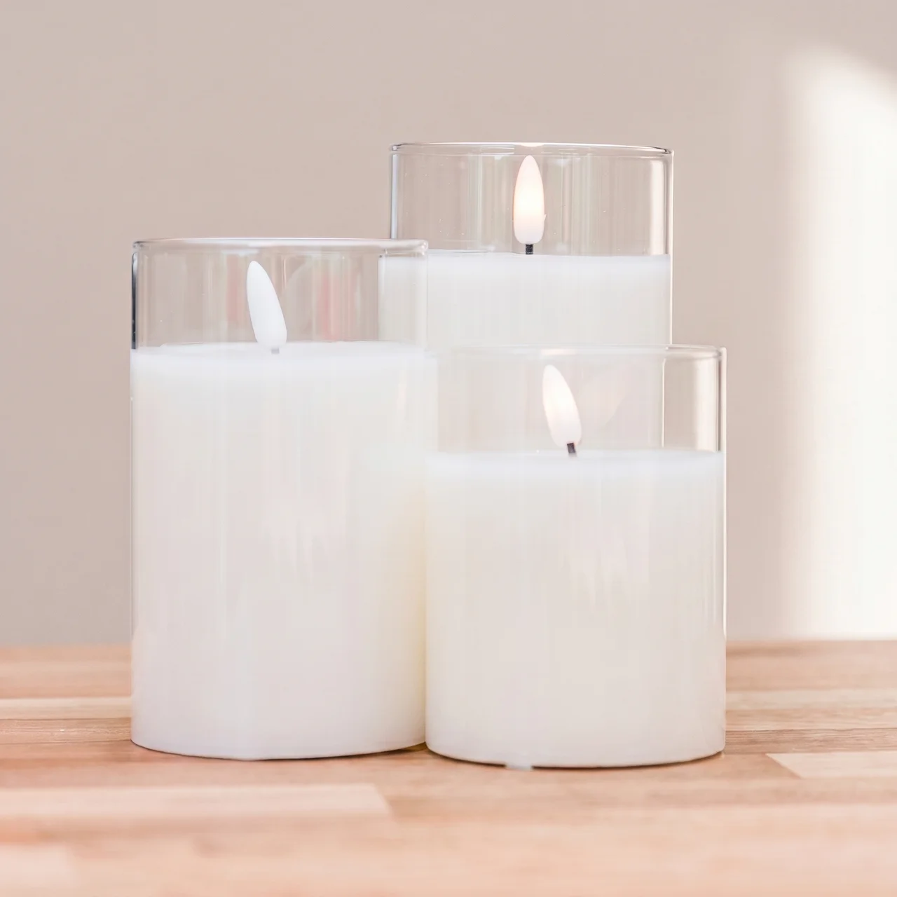 Three white pillar candles in clear glass holders of varying heights, arranged on a light wood surface against a soft beige background.