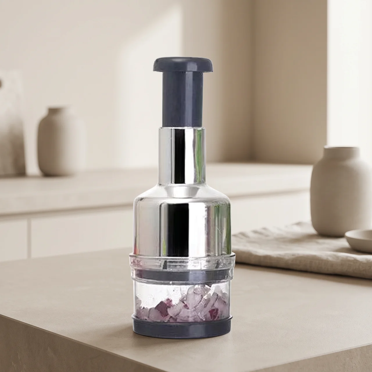 A countertop food chopper with a shiny silver metal body, a dark gray top and base, and a clear lower chamber containing chopped purple onions, set on a beige kitchen counter with blurred ceramic vases in the background.