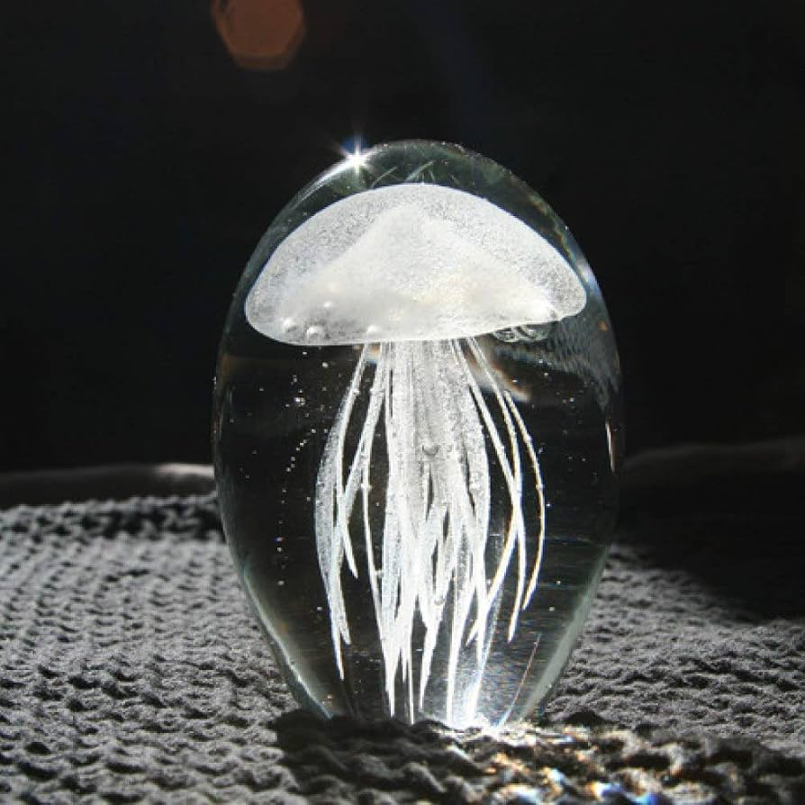 A clear glass or acrylic egg-shaped object containing a white jellyfish design, photographed against a dark background on a textured gray surface.