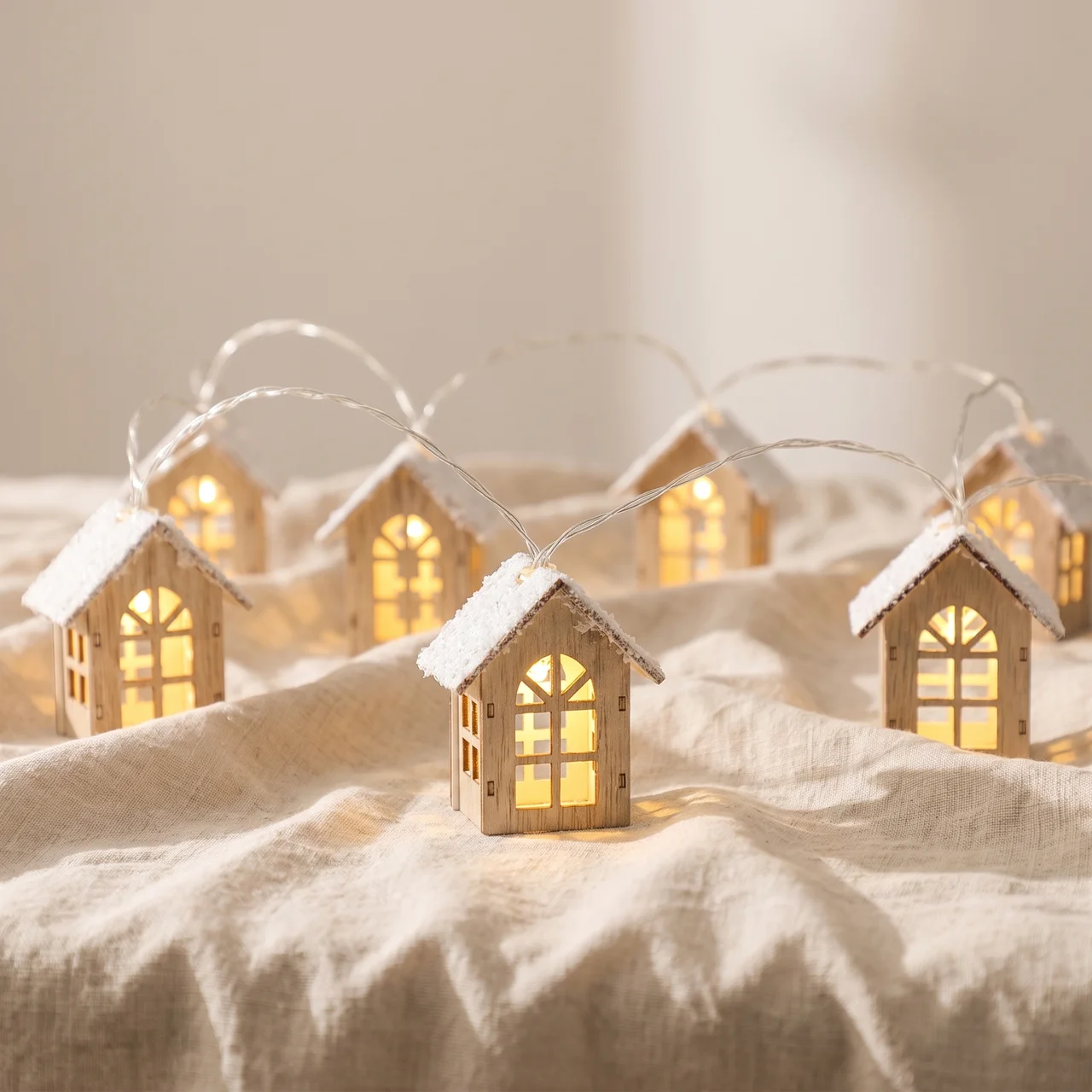 A set of miniature wooden house string lights with glowing warm yellow windows and white faux-snow on the roofs, arranged on a beige linen fabric background.
