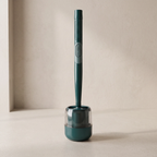A dark teal electric toothbrush with a textured grip and a silver accent band, standing upright in its matching dark teal charging base on a light beige surface against a textured beige wall.