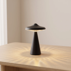 A small black mushroom-shaped table lamp sits on a light wood table, casting a warm radial shadow pattern on the tabletop against a soft beige wall.