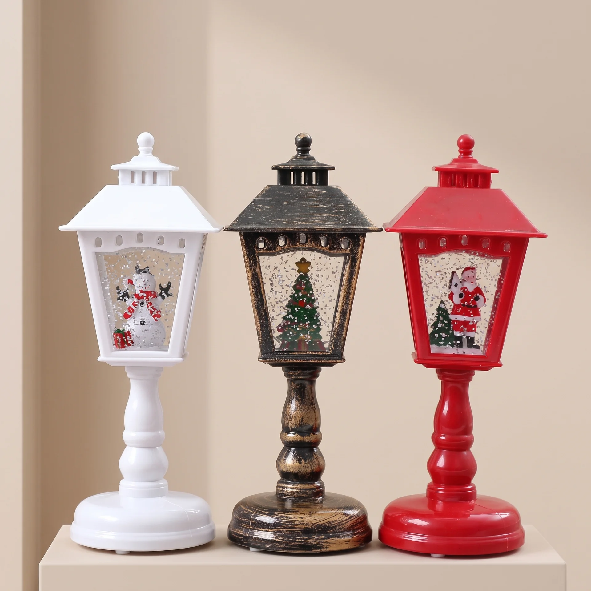 Three decorative table lanterns with holiday designs, shown side by side on a beige pedestal against a beige background. The lanterns are white, dark brown, and red, each with a Santa-themed window display.
