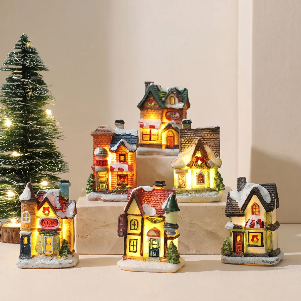A set of miniature Christmas village house figurines with warm glowing windows, displayed on small pedestals with a lit Christmas tree in the background.