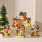 A set of miniature Christmas village house figurines with warm glowing windows, displayed on small pedestals with a lit Christmas tree in the background.