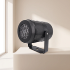 A black cylindrical spotlight with a mounting bracket, shown on a beige pedestal in a minimalist room setting with soft window light.