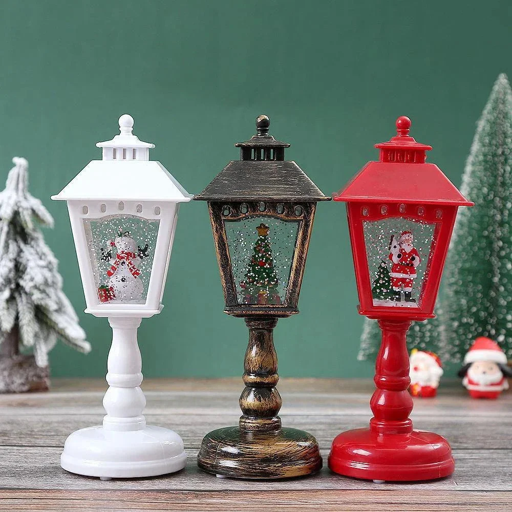 Three small decorative lanterns on stands are displayed on a wooden surface against a green backdrop. The left lantern is white and shaped like a snowman lantern, the center lantern is dark brown with a Christmas tree window, and the right lantern is red with a Santa Claus window.