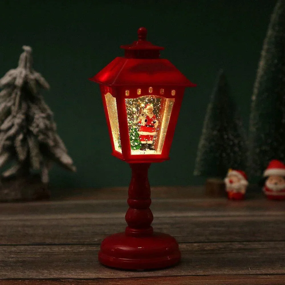 A red table lamp shaped like a vintage street lantern, lit from within to show a miniature Christmas tree scene with Santa Claus. The lamp sits on a rustic wooden surface against a dark green background with blurred Christmas decorations.