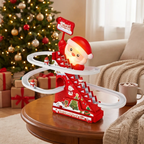 A festive Christmas-themed toy track set on a wooden table in a cozy living room. The red and white toy features a Santa Claus figure, winding white tracks, small decorative hills, and a sign that reads 'Merry Christmas.