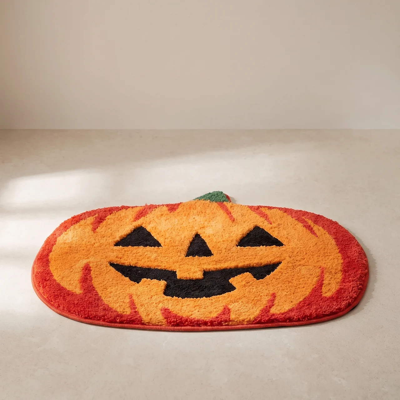 A Halloween-themed plush bath mat shaped like a jack-o'-lantern pumpkin, featuring a bright orange face with black stitched details and a red border, set against a neutral beige background.