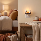 A cozy bedroom scene showing a wall-mounted night light with two white ghost figures around a small flame, lit in a warm beige room with a bed, table, blanket, and basket visible.