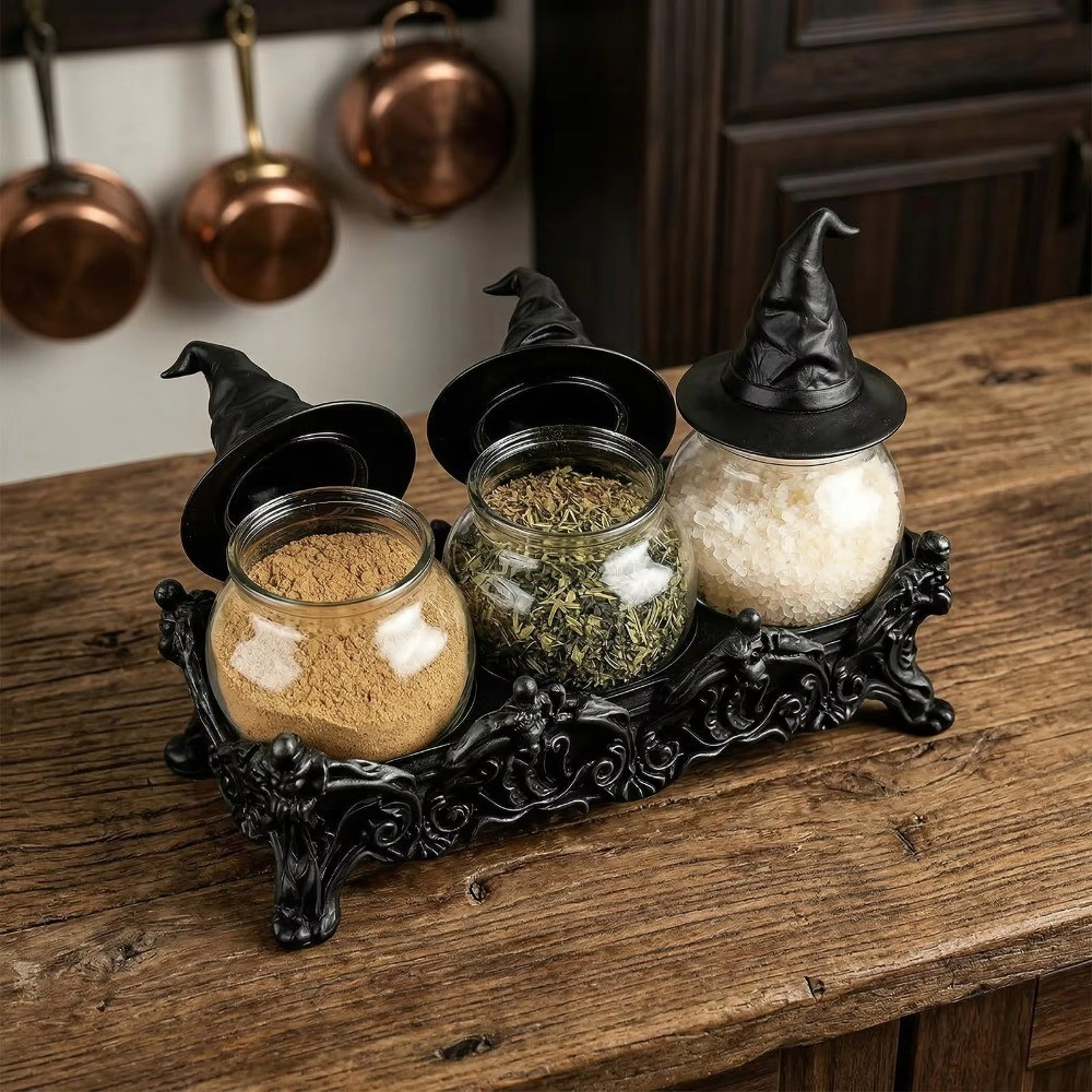 Three glass spice jars with black witch hat lids sit on an ornate black decorative tray on a rustic wooden table. Copper pots hang in the background.