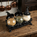 Three glass spice jars with black witch hat lids sit on an ornate black decorative tray on a rustic wooden table. Copper pots hang in the background.
