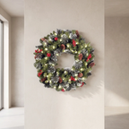 A pre-lit Christmas wreath with green pine branches, white snow dusting, red berries, and pinecones, hanging on a beige wall.