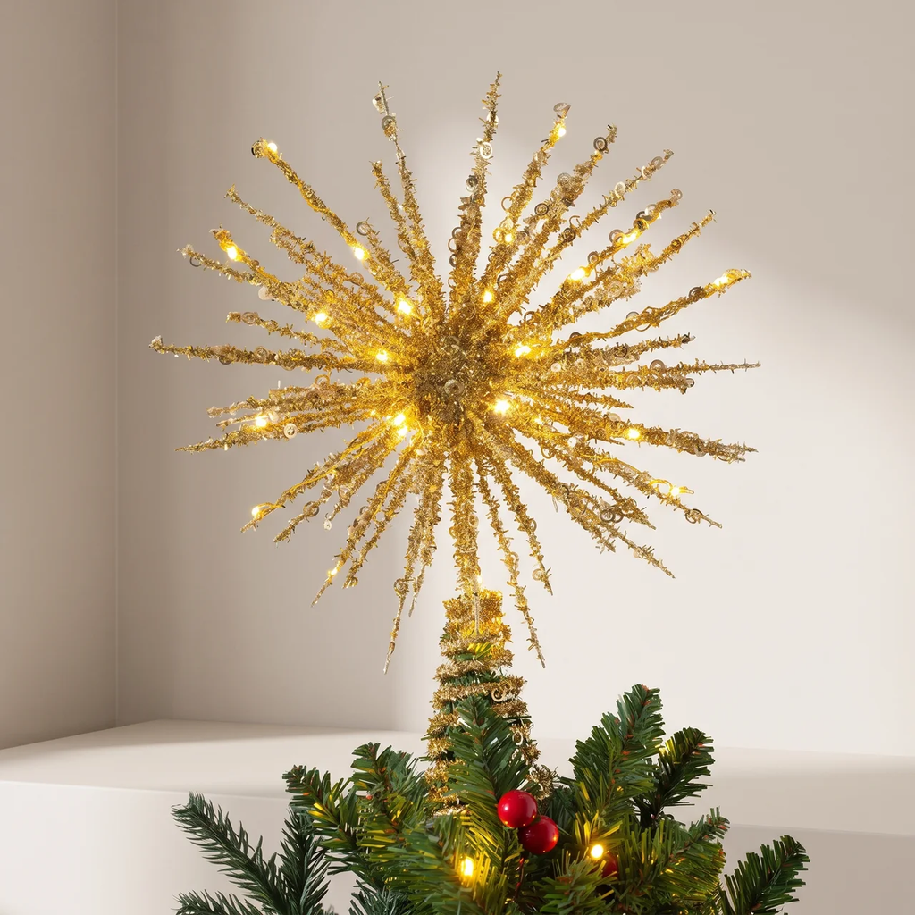 A gold glittery starburst Christmas tree topper with warm white lights, mounted on a green artificial pine tree with small red ornaments, set against a plain off-white wall.