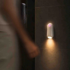 A white cylindrical wall-mounted light fixture glowing with warm light on a dark tiled wall, with a blurred person partially visible in the foreground.
