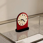 A modern arch-shaped desk clock with a red body, cream dial face, and black hands, displayed on a glass tabletop with a mesh surface.