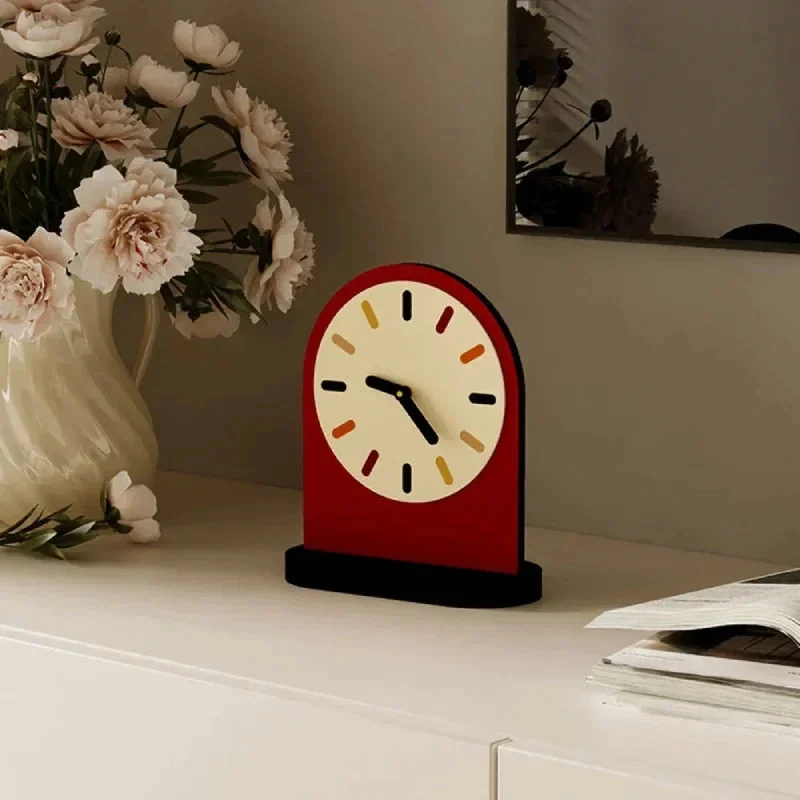 A retro-style arched desk clock with a red body, cream dial, and black hands, placed on a white tabletop beside a vase of pale flowers and an open magazine.