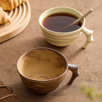 Two ceramic mugs with handles, one containing a dark liquid and a spoon, on a textured surface.