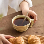 Person holding a cup of coffee with a spoon, next to two croissants on a wooden board.