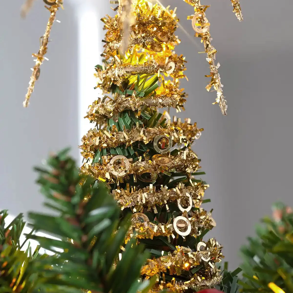 A close-up of a decorated artificial Christmas tree with green needles, wrapped in shiny gold tinsel, and adorned with small gold ornaments against a soft gray background.