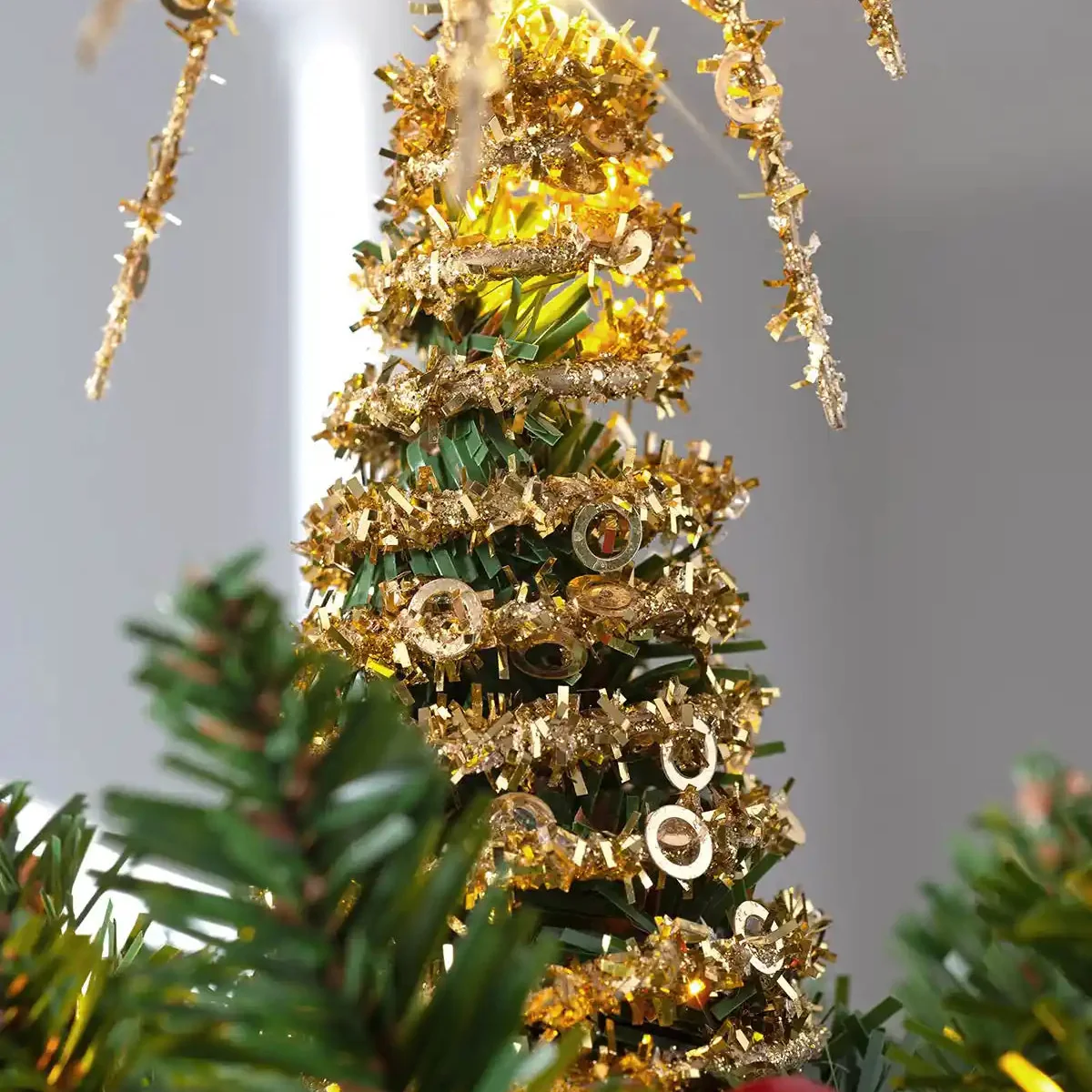 A close-up of a decorated artificial Christmas tree with green needles, wrapped in shiny gold tinsel, and adorned with small gold ornaments against a soft gray background.