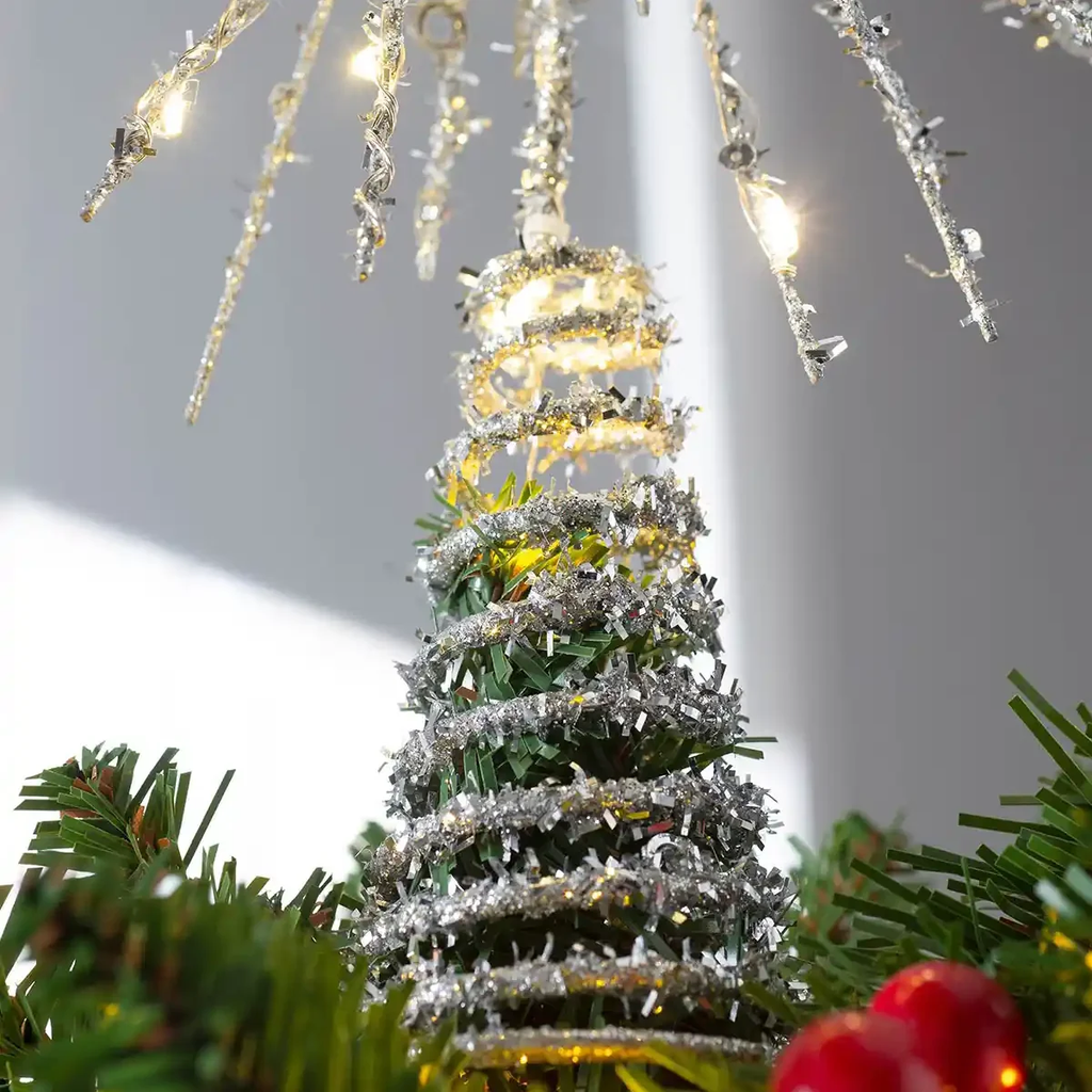 A close-up of a silver tinsel Christmas tree ornament with warm white string lights, surrounded by green pine needles and red ornaments in the foreground.