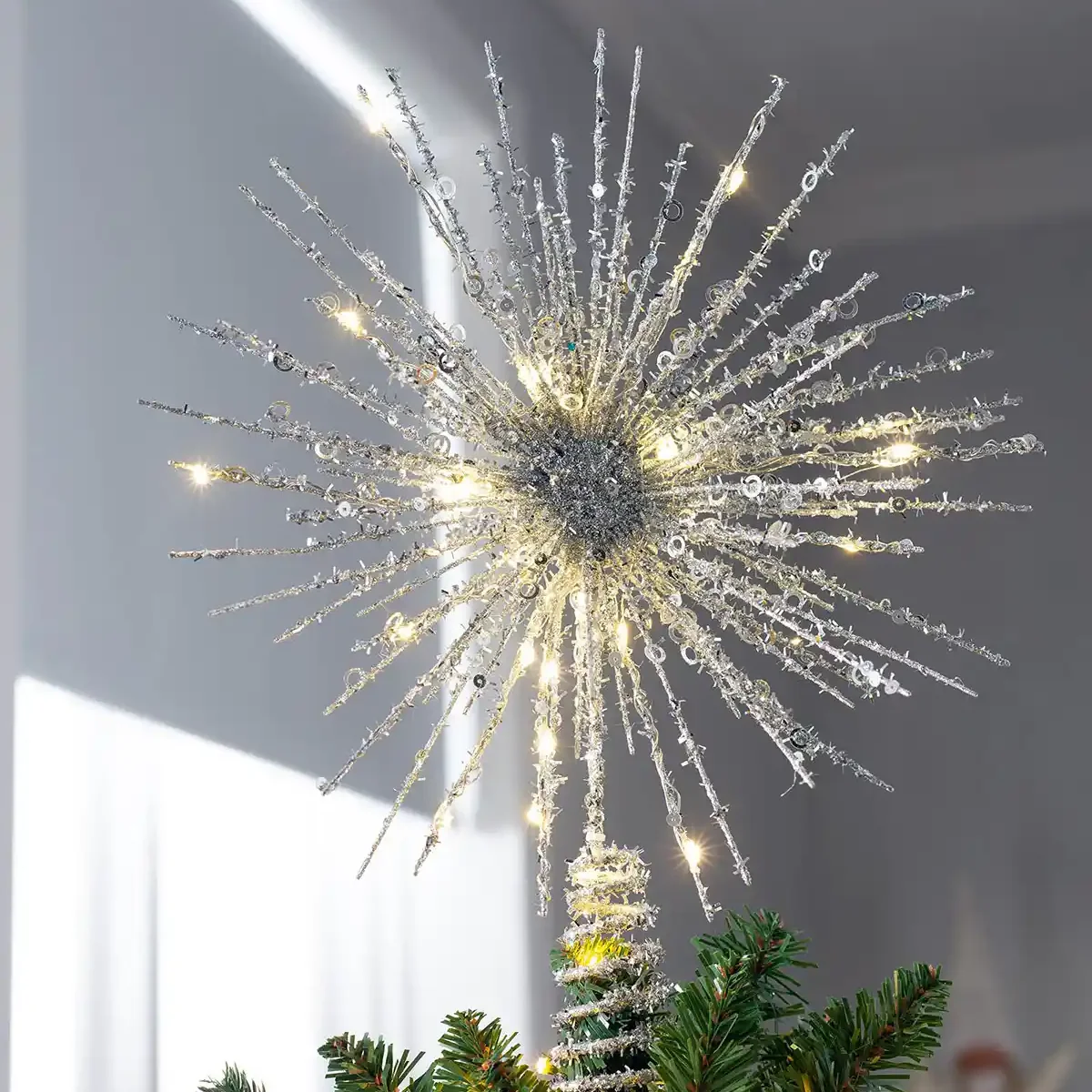 A sparkling star-shaped Christmas tree topper with radiating silver branches, clear crystal-like accents, and warm white LED lights, mounted on a decorated tree with green pine needles against a soft gray indoor background.