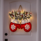 A festive Christmas wreath hanging on a white front door. The wreath features a red stocking-shaped lower section with white snowflake details, a white faux-fur trim, and a top section made of green pine branches, white snowflakes, pinecones, and warm white string lights with red berry accents.