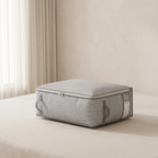 A rectangular gray fabric storage bin with a zippered lid and clear side panels sits on a bed in a softly lit bedroom.