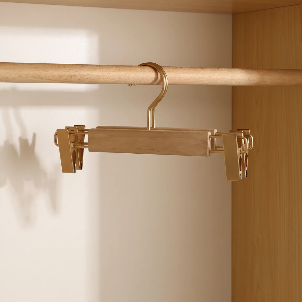 A wooden hanger with gold-toned metal clips and a gold hook, shown hanging from a wooden closet rod inside a closet.