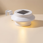 A white solar-powered outdoor light fixture with a circular top panel containing a dark solar cell and a side mounting bracket, shown lit on a warm beige background.