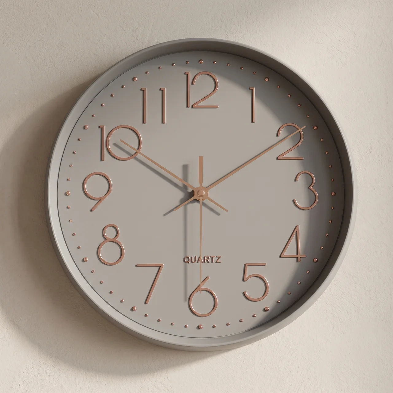 A minimalist round wall clock with a light gray face and frame, rose gold numerals and hands, and a subtle drop shadow on a textured off-white wall.
