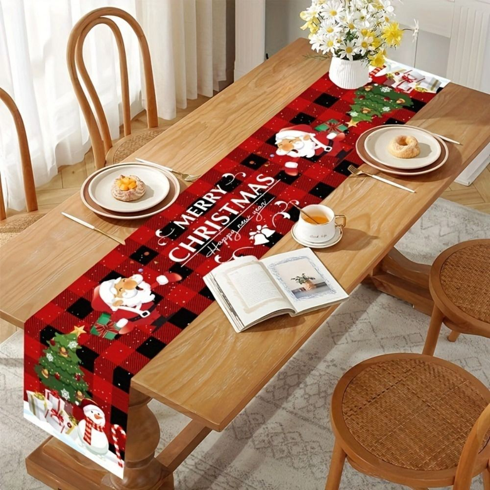 A red and black plaid Christmas table runner with Santa Claus, trees, and 'MERRY CHRISTMAS' text, styled on a wooden dining table with plates, cups, and chairs.