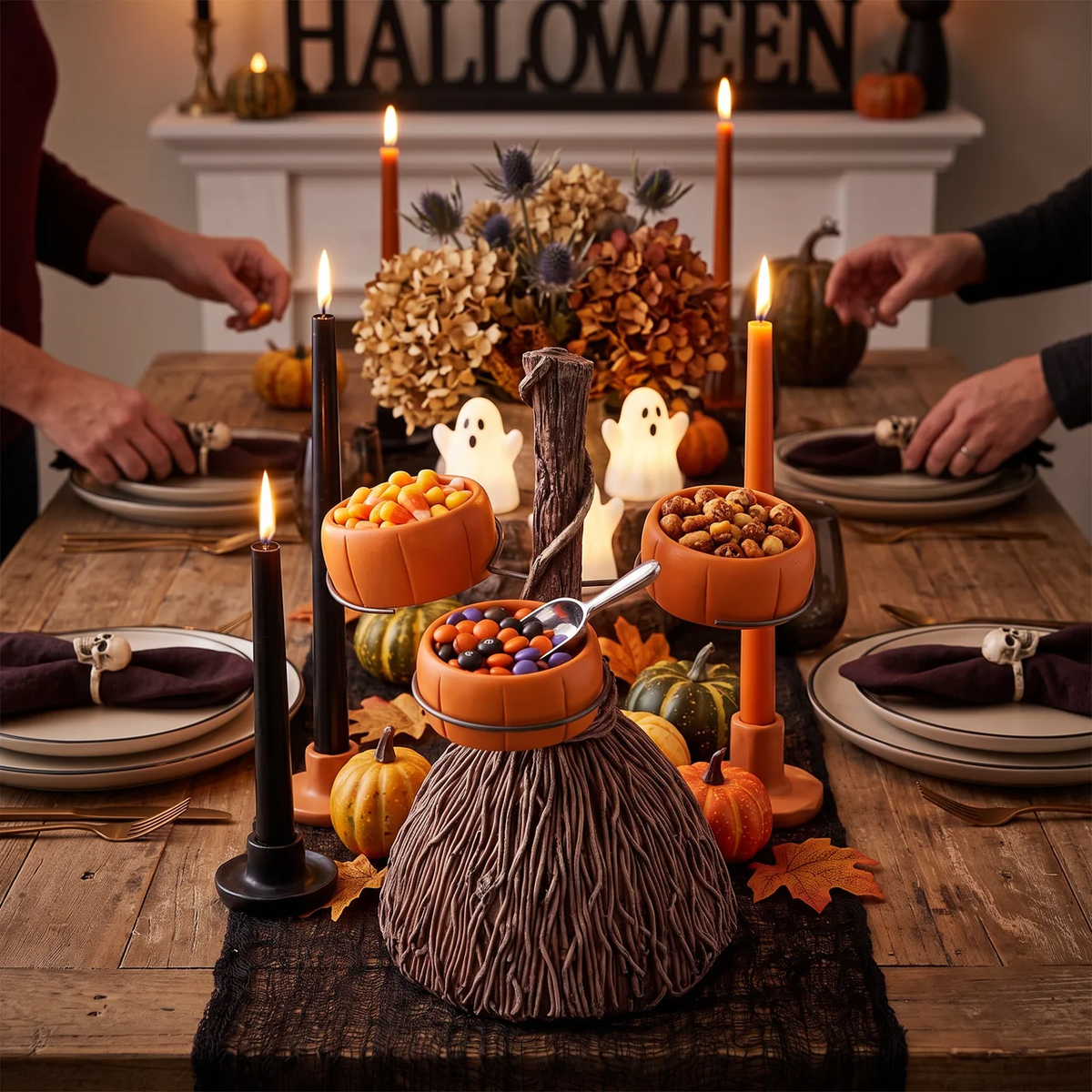 A styled Halloween table centerpiece with a brown twig-like pumpkin base holding orange pumpkin-shaped bowls of candy and nuts, surrounded by black candles, small pumpkins, white ghost decorations, and place settings. Partial human arms and hands are visible at the table edges in a warmly lit dining setting.
