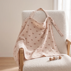 A soft muslin baby blanket in a dusty pink color with a repeating brown crescent moon pattern, draped over a cream-colored textured armchair.